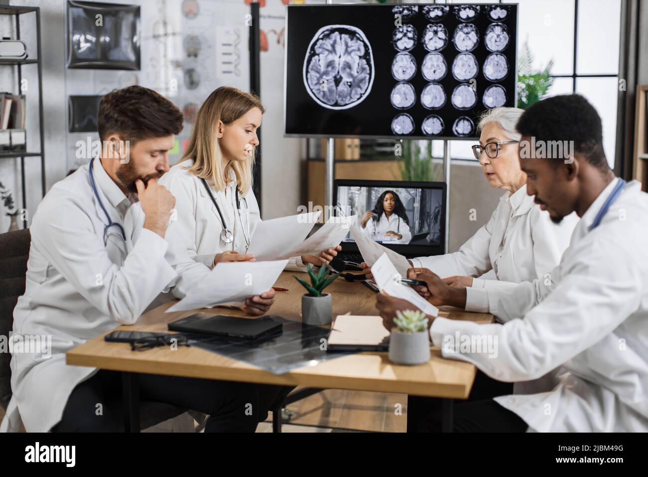 Gruppo multiculturale di quattro ricercatori medici che hanno una formazione online in laboratorio con monitor sullo sfondo che mostra l'immagine MRI cerebrale. Colleghi che condividono idee mentre tengono l'elenco della storia del paziente. Foto Stock