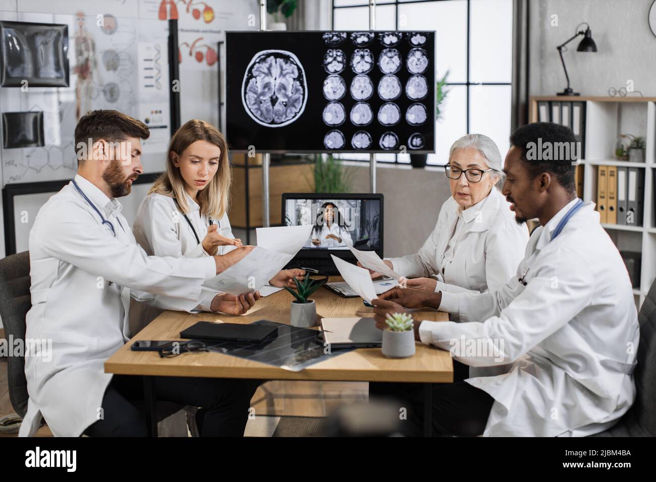 Gruppo multiculturale di quattro ricercatori medici che hanno una formazione online in laboratorio con monitor sullo sfondo che mostra l'immagine MRI cerebrale. Colleghi che condividono idee mentre tengono l'elenco della storia del paziente. Foto Stock