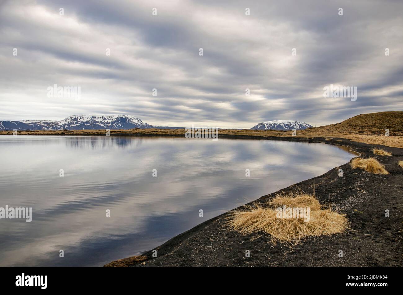 Vista da una spiaggia con sabbia nera e ghiaia attraverso un lago riflettente nella zona di Myvatn in Islanda verso una catena montuosa innevata sotto un dramma Foto Stock