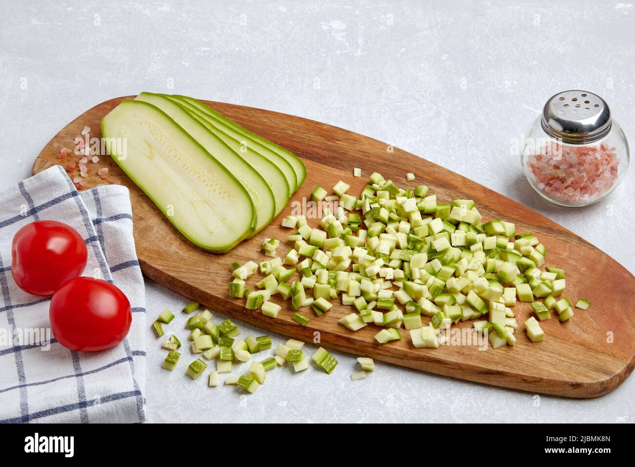 Zucchine a dadini tagliate a fette su un tagliere di legno, pomodori, sale rosa su un tavolo chiaro di cemento Foto Stock