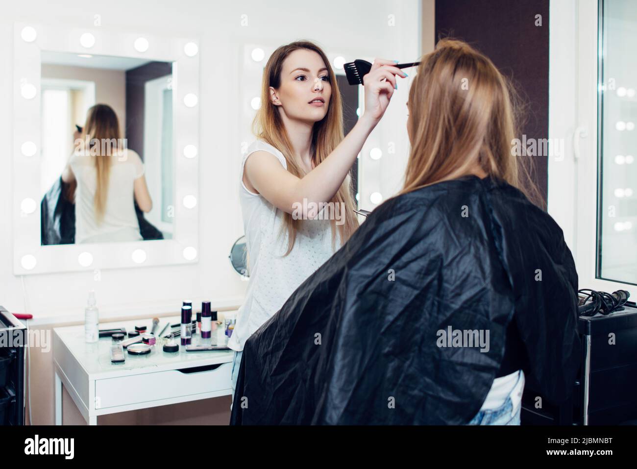 Hairstylist che separa i capelli lunghi e giusti della giovane cliente femminile seduta in cape al salone di parrucchiere. Foto Stock