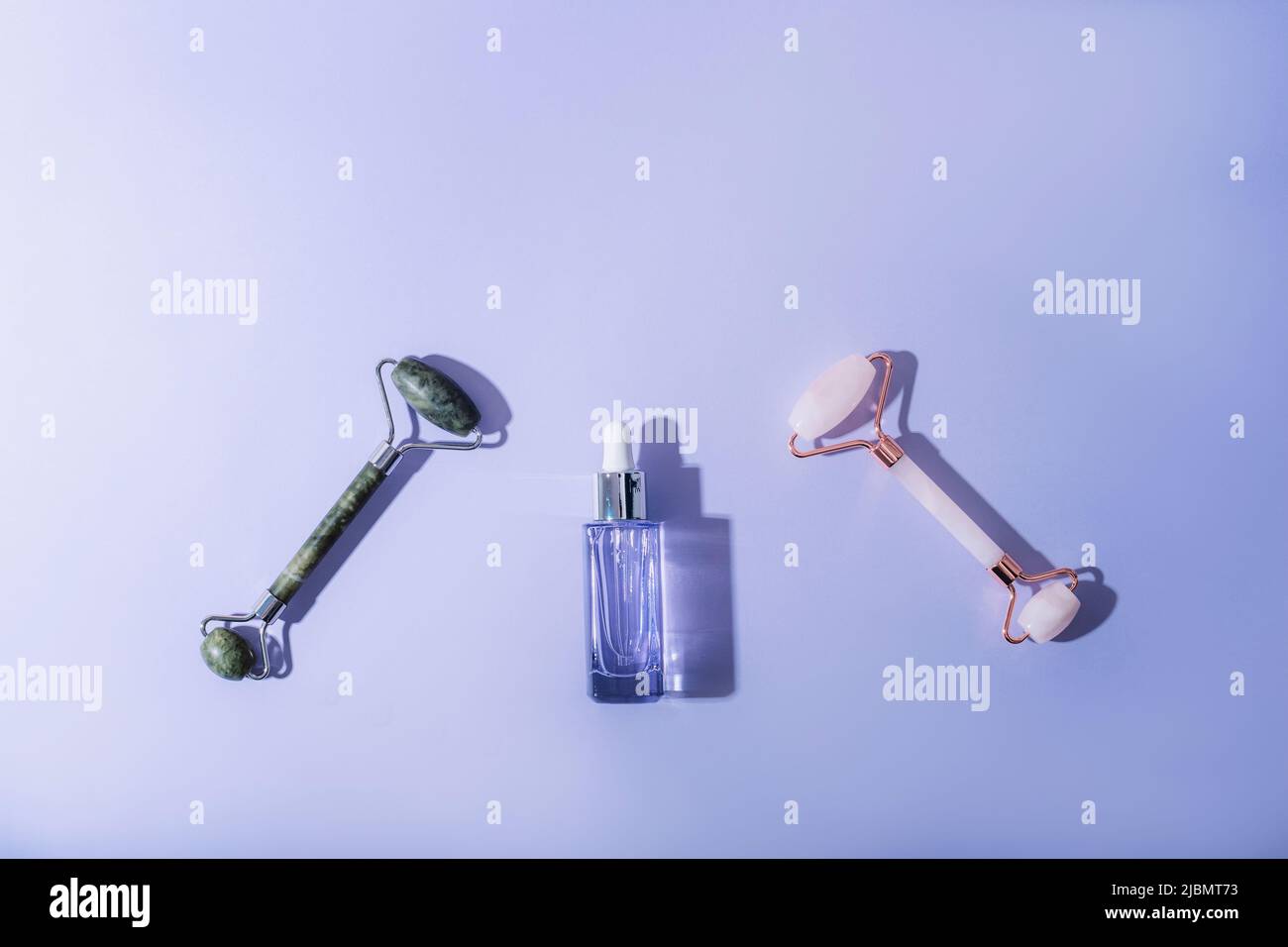 Bottiglia di siero e due rulli facciali su sfondo viola con ombre dure. Cura dell'auto, bellezza, spa e concetto di benessere. Vista dall'alto, disposizione piatta, spazio di copia Foto Stock