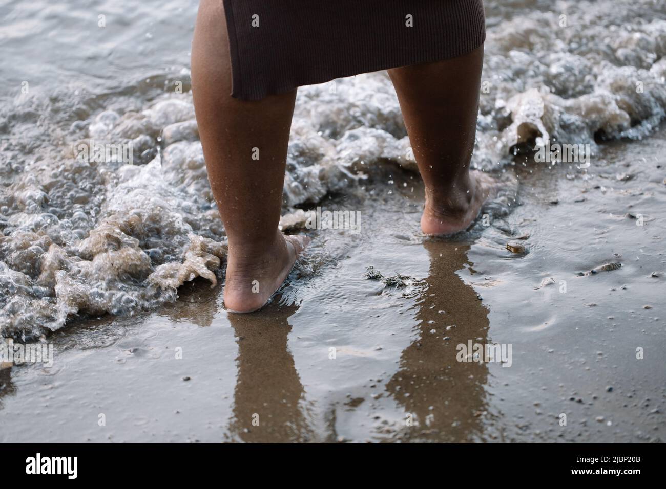Cropped pelle scura a piedi nudi donna gambe camminare attraverso la costa del mare, spiaggia, schiuma lavaggio. Multiculturale, vacanze estive Foto Stock