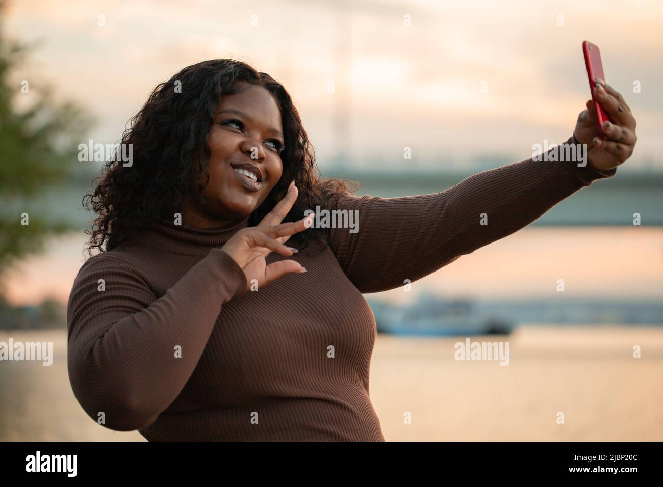 Sorridente e gioiosa bella afroamericana donna in abito elegante prendendo selfie da smartphone. Vacanze estive vicino al mare Foto Stock