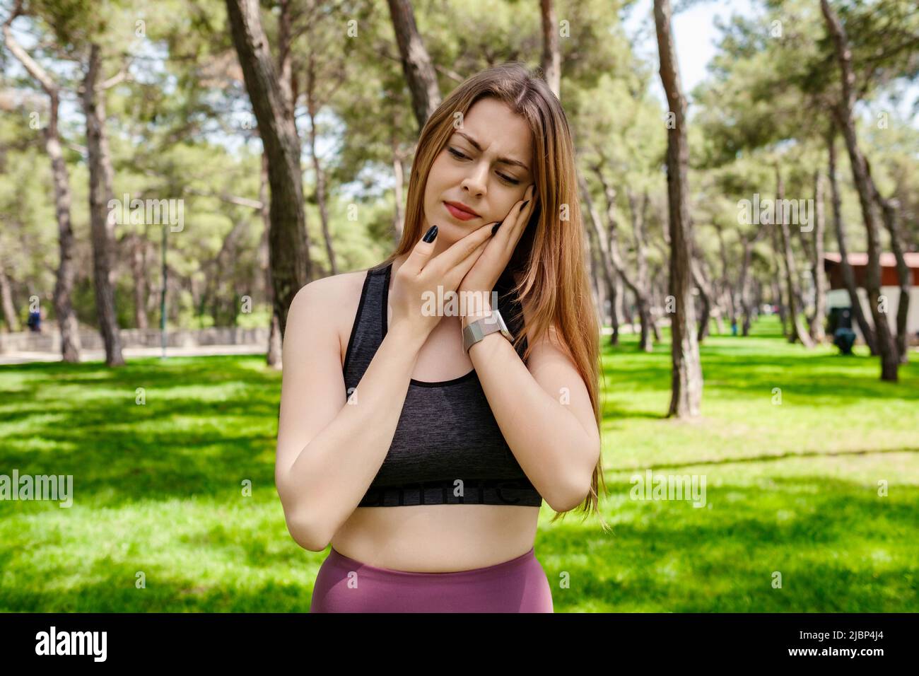 Ritratto di donna allegra indossando un reggiseno sportivo nero in piedi sul parco della città, all'aperto toccando la bocca con la mano con espressione dolorosa a causa di denti Foto Stock