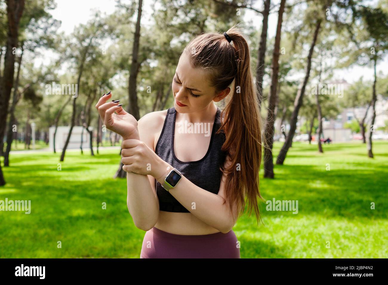 Ritratto di giovane donna bruna con reggiseno sportivo in piedi sul parco della città, all'aperto tenendo il suo polso doloroso. Dolore alle mani e alle dita, ar Foto Stock