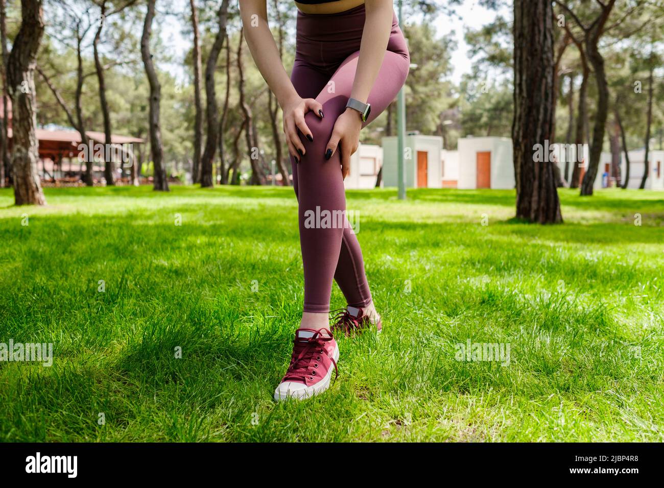 Donna sportiva bruna con reggiseno sportivo nero in piedi sul parco cittadino, all'aperto toccando il ginocchio ferito con le mani. Gamba ferita, stile di vita sano e. Foto Stock