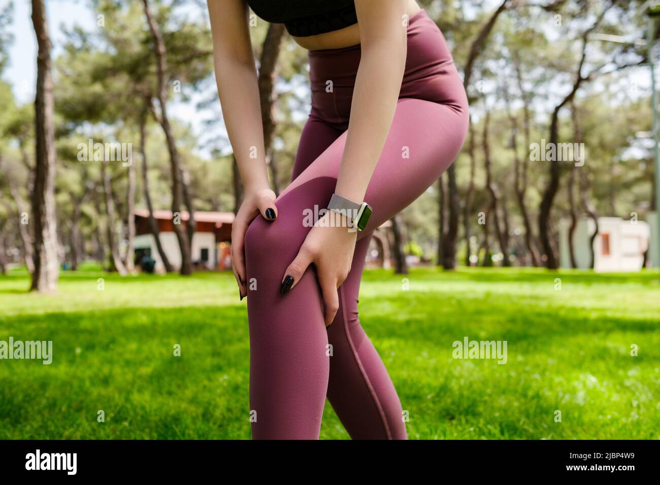 Bella donna bruna con reggiseno sportivo in piedi sul parco cittadino, all'aperto toccando il ginocchio ferito con le mani. Gamba ferita, stile di vita sano e sport Foto Stock
