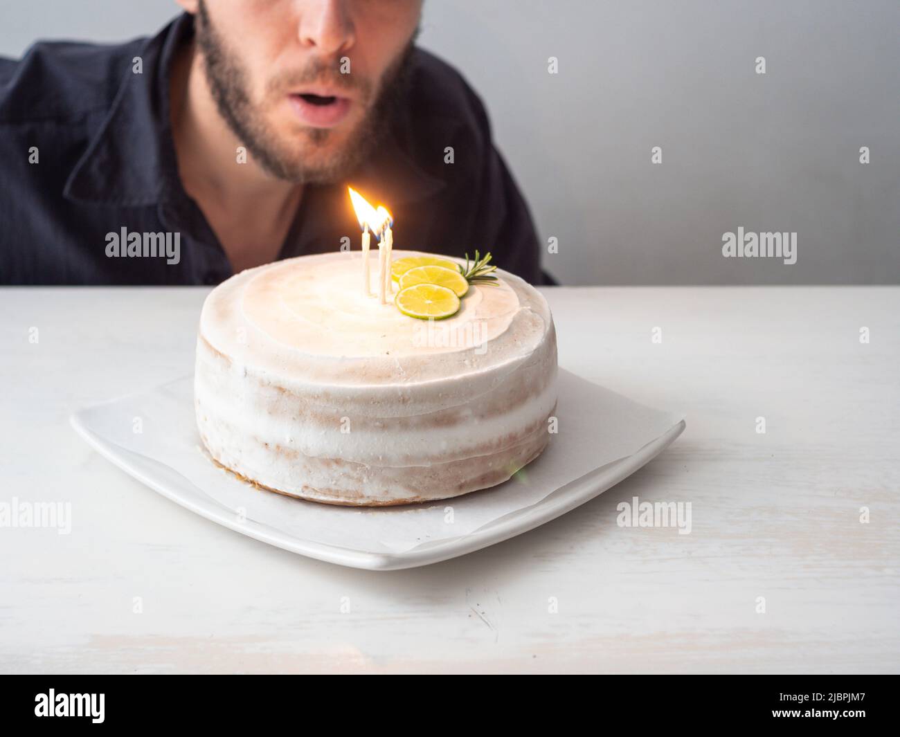 L'uomo bianco soffia le candele su una torta di compleanno Vegan con mandorle, cocco e base di limone su un tavolo bianco e uno sfondo bianco Foto Stock