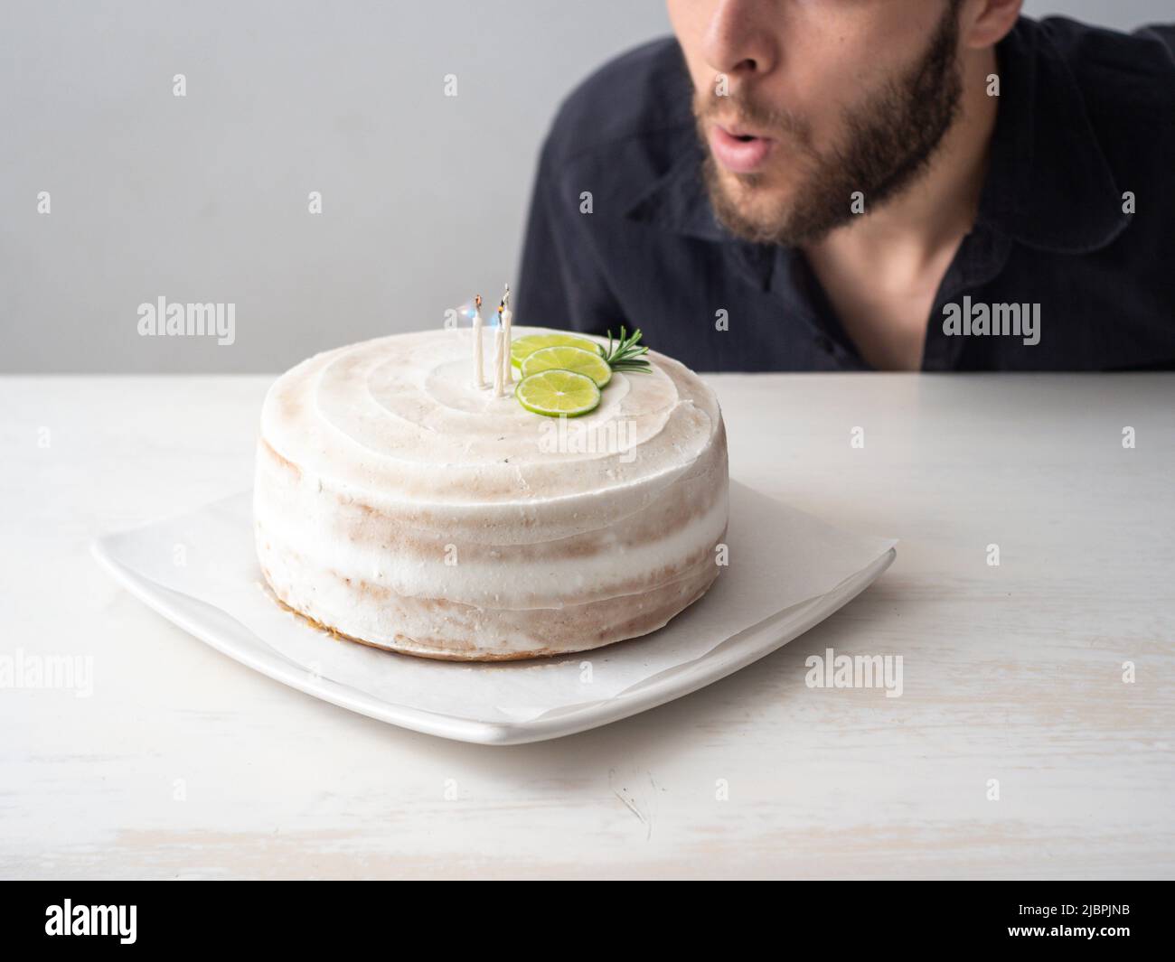 L'uomo bianco soffia le candele su una torta di compleanno Vegan con mandorle, cocco e base di limone su un tavolo bianco e uno sfondo bianco Foto Stock