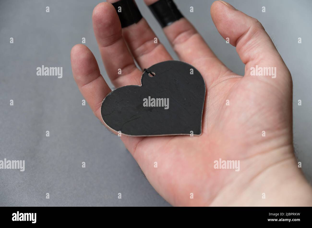 Il cuore nero si trova nel palmo di un uomo. Figura in carta nera a forma di cuore con filo nero. La mano di un uomo di mezza età contro un backgroun grigio Foto Stock