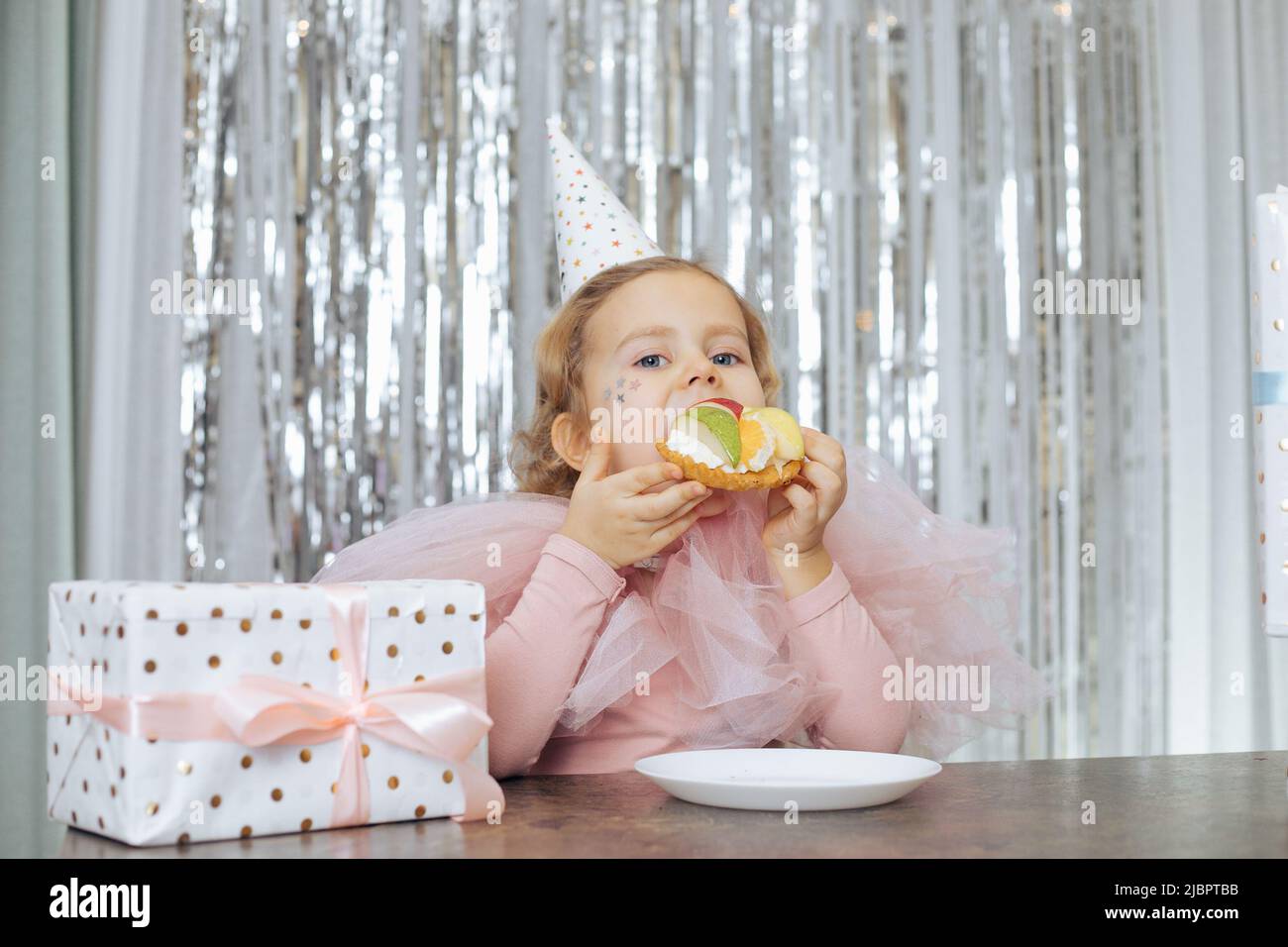 Affascinante, fesivamente vestito rosso-capelli bambino 4-5 anni gode torta deliziosa per il suo compleanno, seduto al tavolo in caffetteria, accanto alla scatola del regalo. Bambini Foto Stock