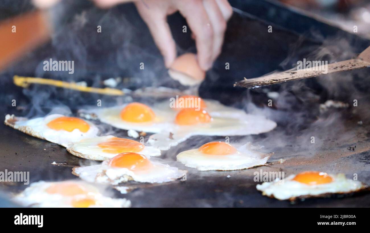 Un certo numero di uova crude fresche che sfrigolano via su un caldo nero cuoco cuoco del barbecue fumante. Un uovo è incrinato sullo sfondo per essere cucinato. Uova sul barbecue s Foto Stock
