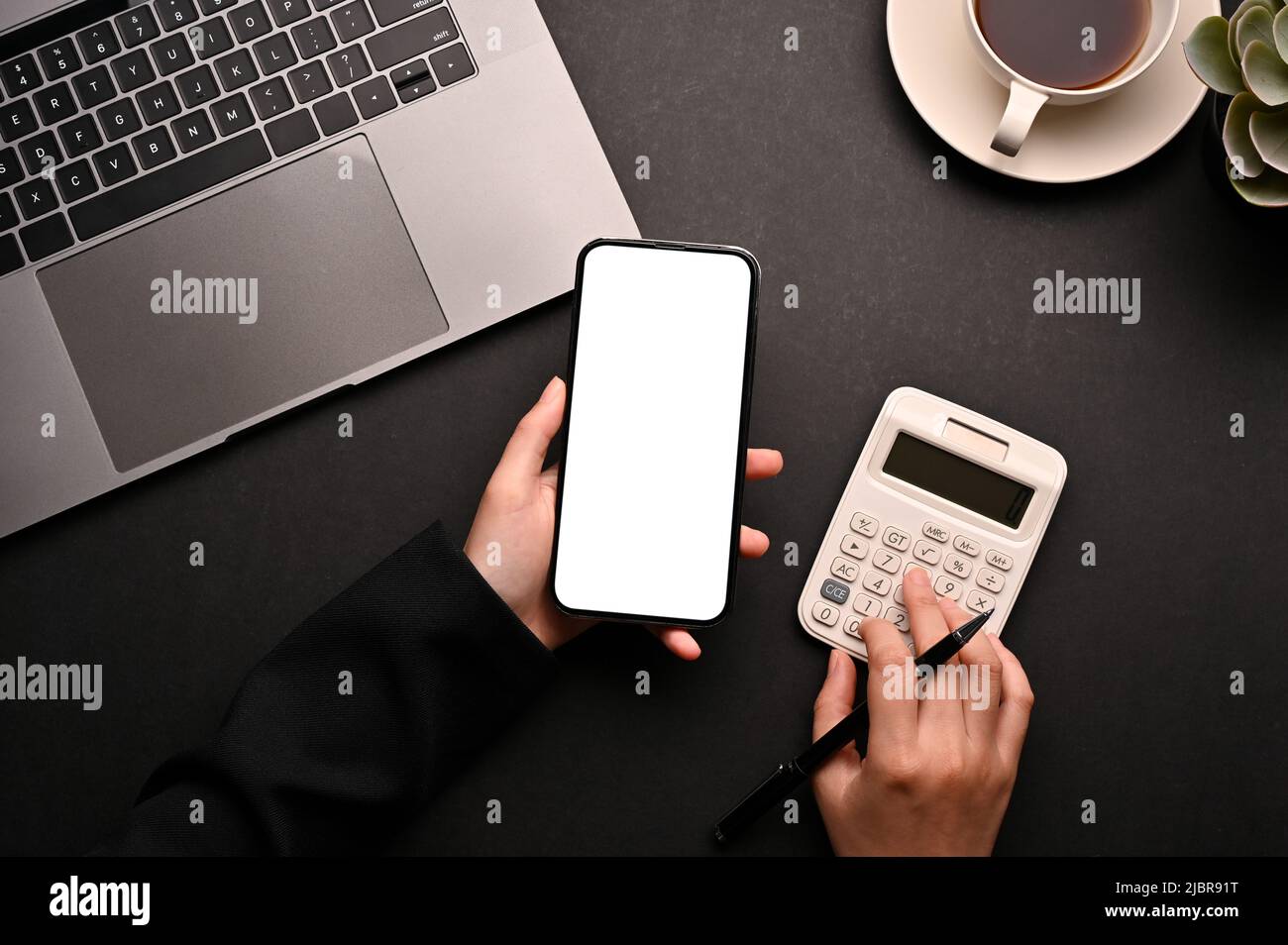 Una donna che calcola le spese mensili utilizzando smartphone e calcolatrice nella sua moderna scrivania da ufficio nera. Vista dall'alto Foto Stock