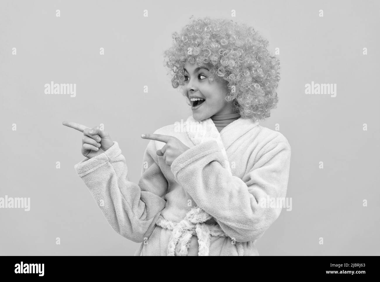 bambino felice in fantasia parrucca arancione capelli indossare home accappatoio puntando dito su spazio copia, pubblicità Foto Stock