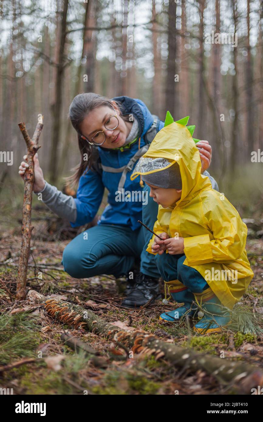 Mamma e bambino camminano nella foresta dopo la pioggia in impermeabile insieme, guardando i funghi su un albero caduto e parlando Foto Stock