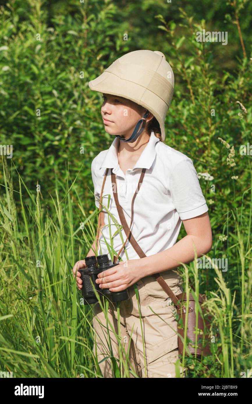 La ragazza che indossa il casco di pith si alza nell'erba che tiene binocoli osservando la natura estiva. Concetto di scoperta e avventure Foto Stock