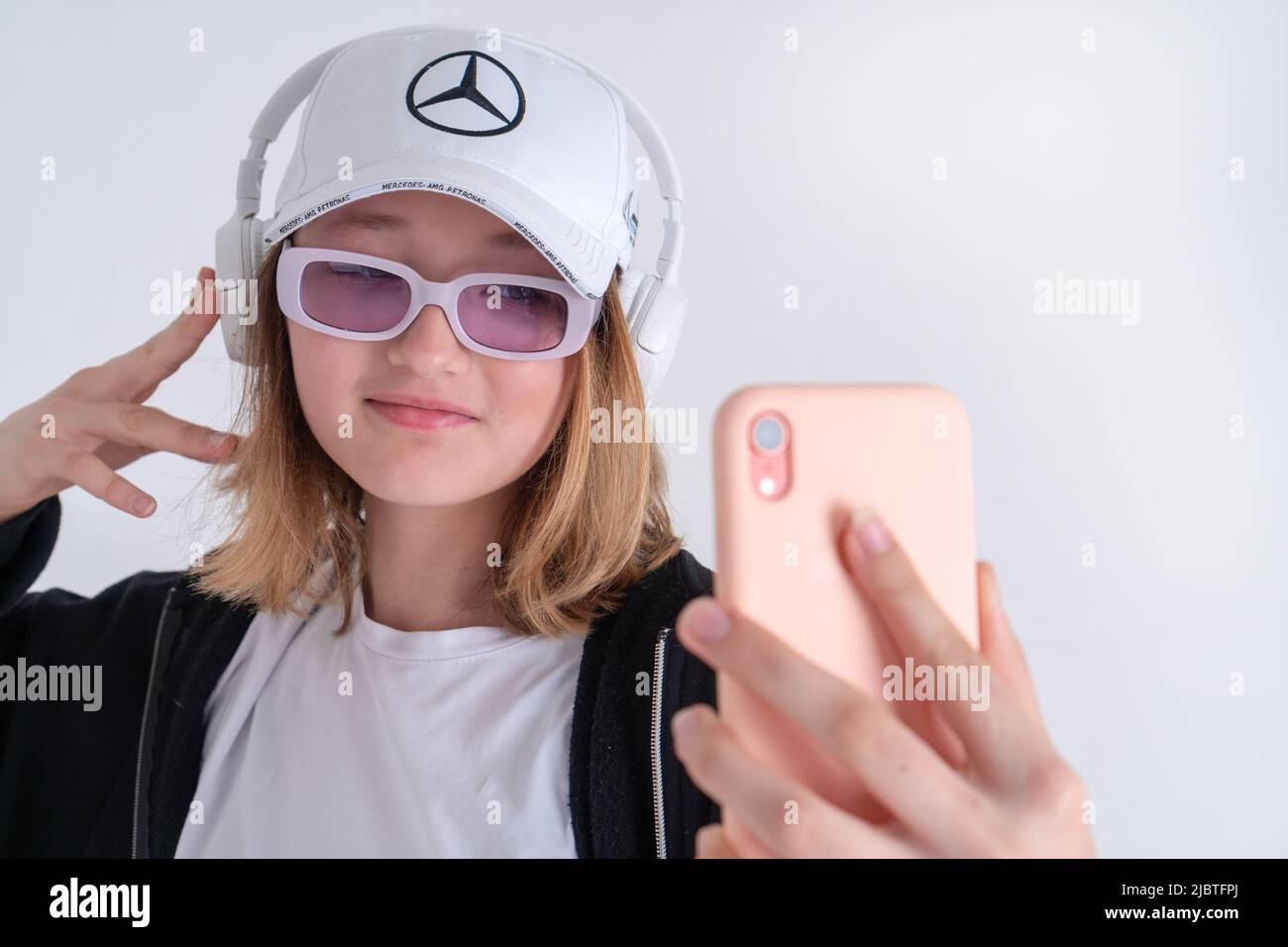 Ritratto di un'adorabile ragazza teen elegante in un cappello con il logo della squadra Mercedes in occhiali da sole. Cuffie bianche JBL scatta un video e scatta una foto Foto Stock