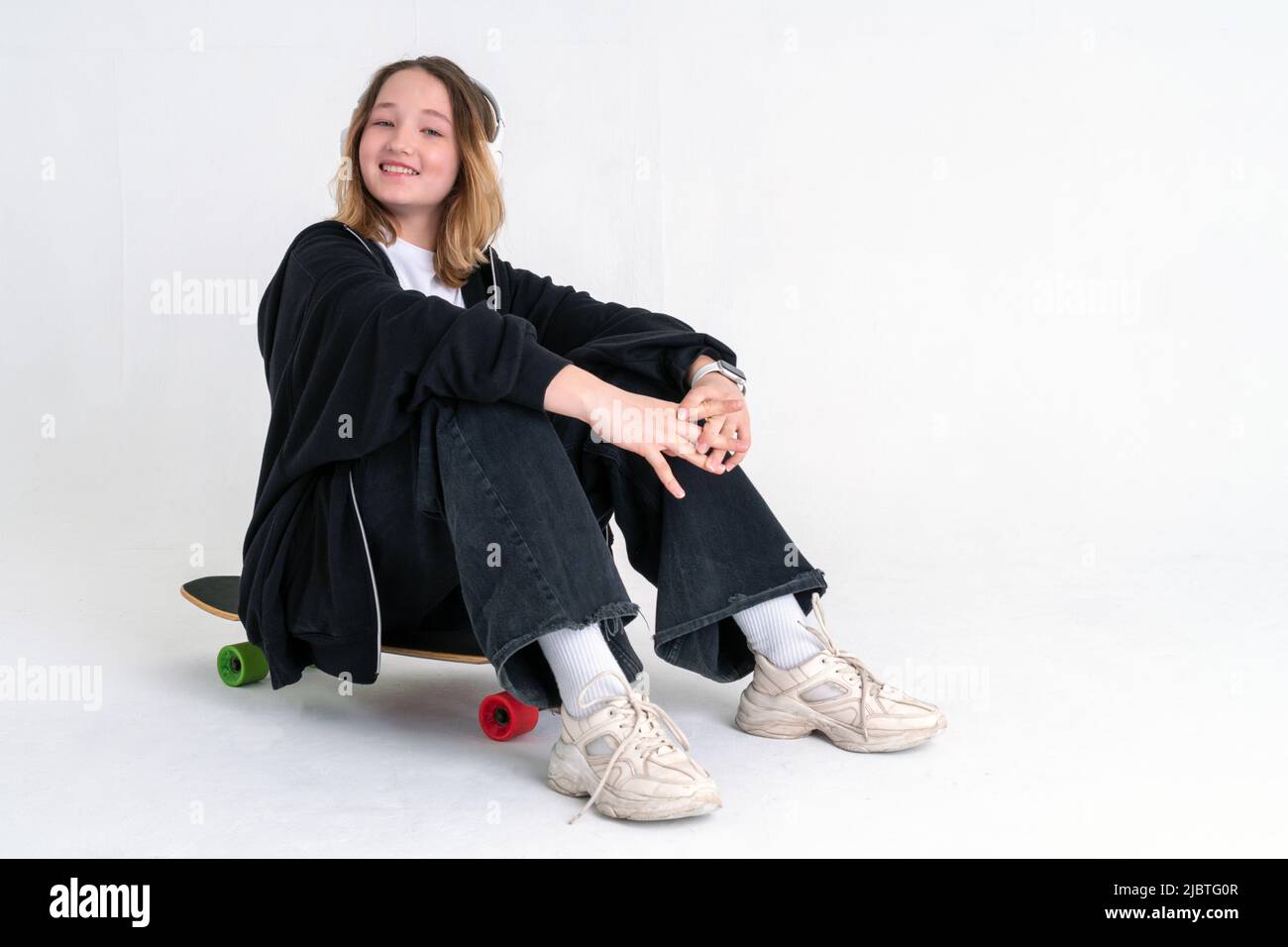 Un'elegante ragazza adolescente con un acconciatura in un felpa nera con cappuccio e jeans neri si siede su una tavola lunga in cuffie e sorride su uno sfondo bianco. Spac Foto Stock