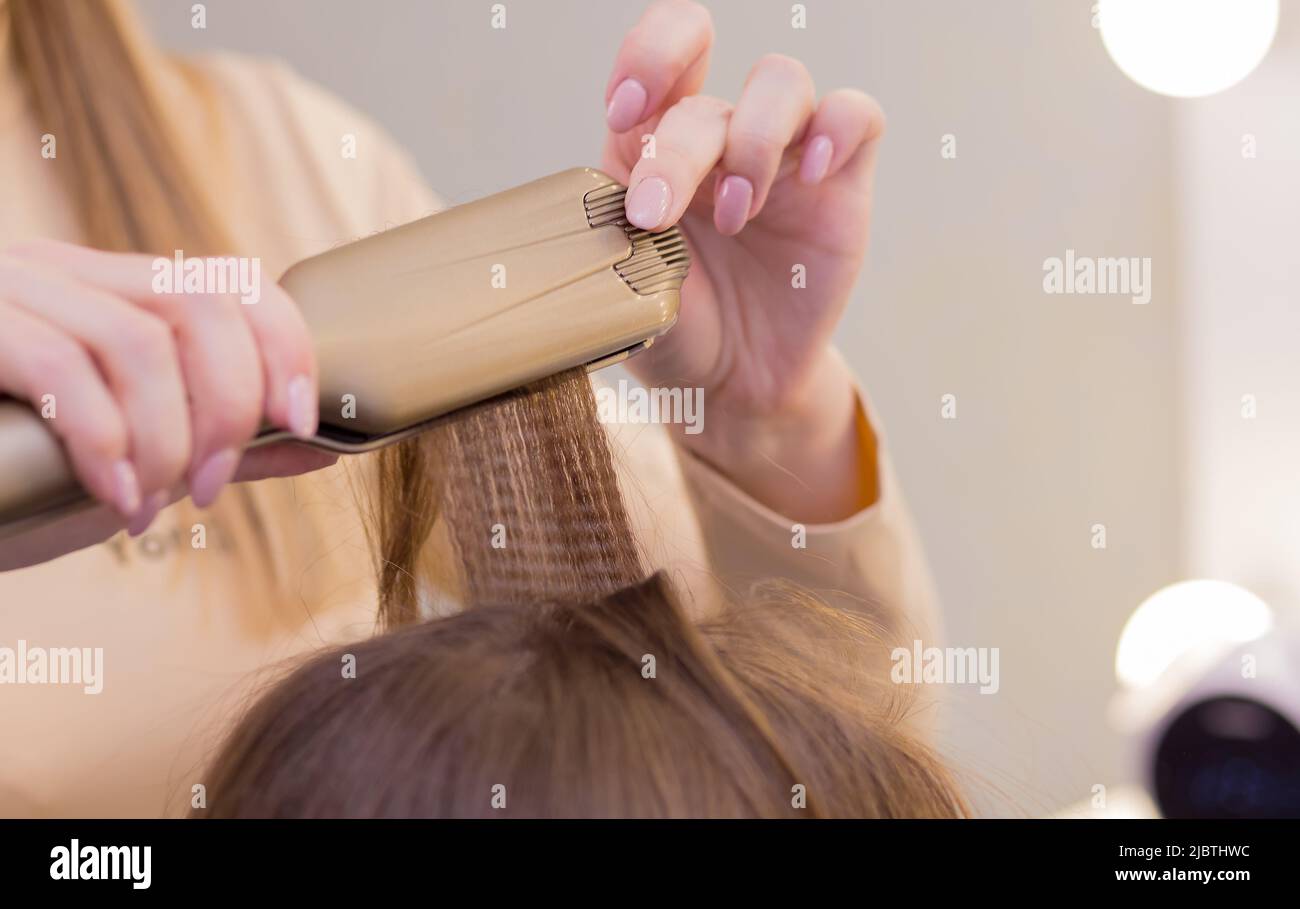 Il primo piano delle mani femminili con un ferro arricciante raddrizza una ciocca di capelli. Il parrucchiere crea un'acconciatura per una giovane donna. Barbiere, concetto di business. Salone di bellezza, cura dei capelli. Foto Stock