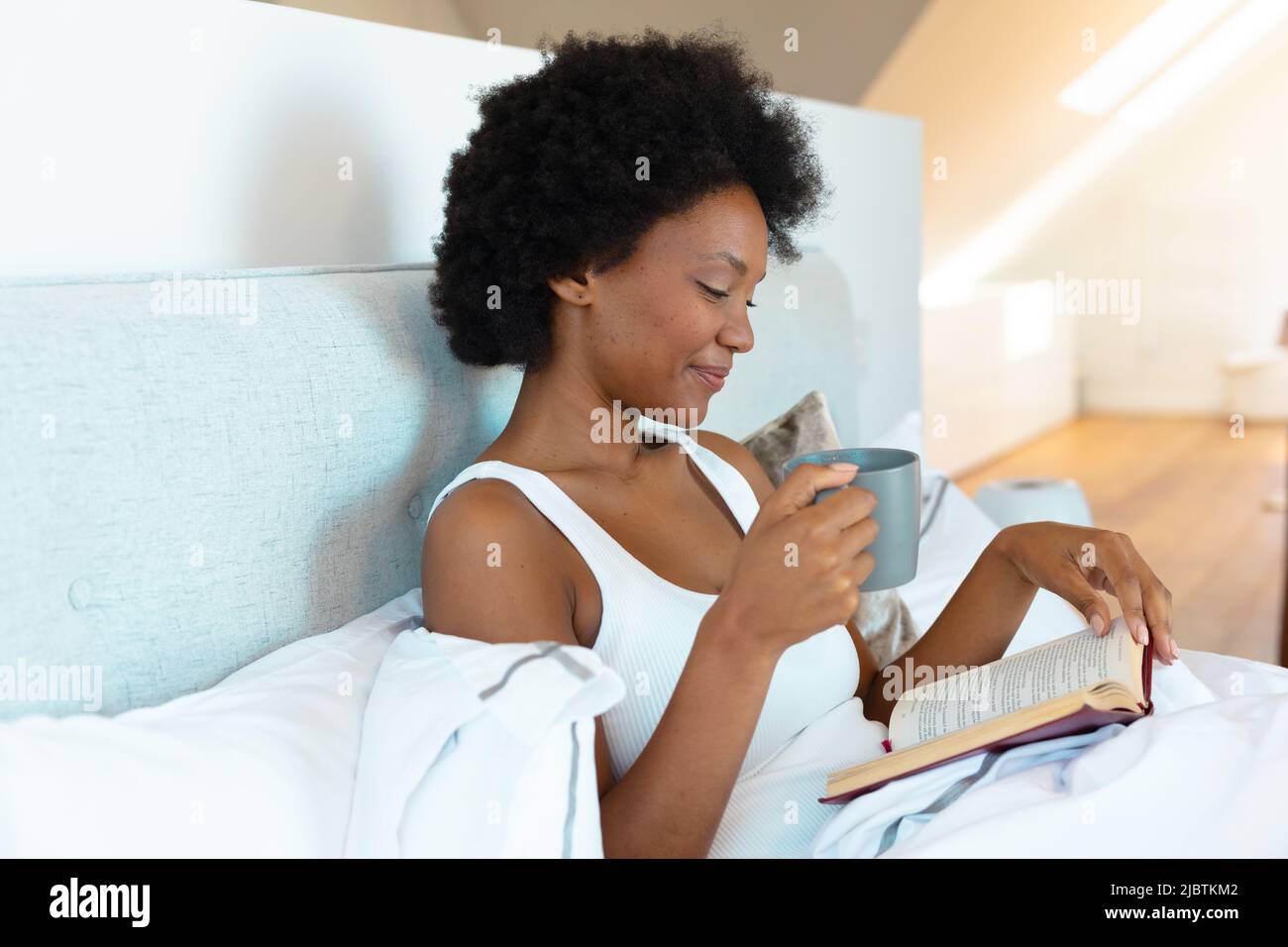 Giovane donna afroamericana che tiene una tazza di caffè leggendo un libro nel letto di casa Foto Stock