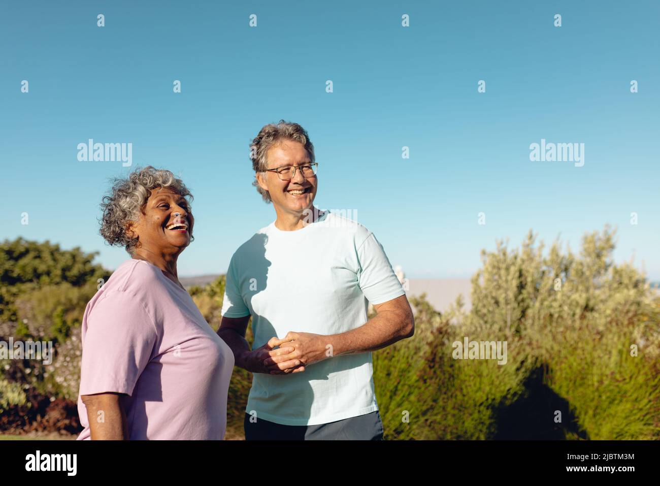 Multirazziale amici anziani felici in piedi contro le piante contro il cielo blu chiaro durante la giornata di sole Foto Stock
