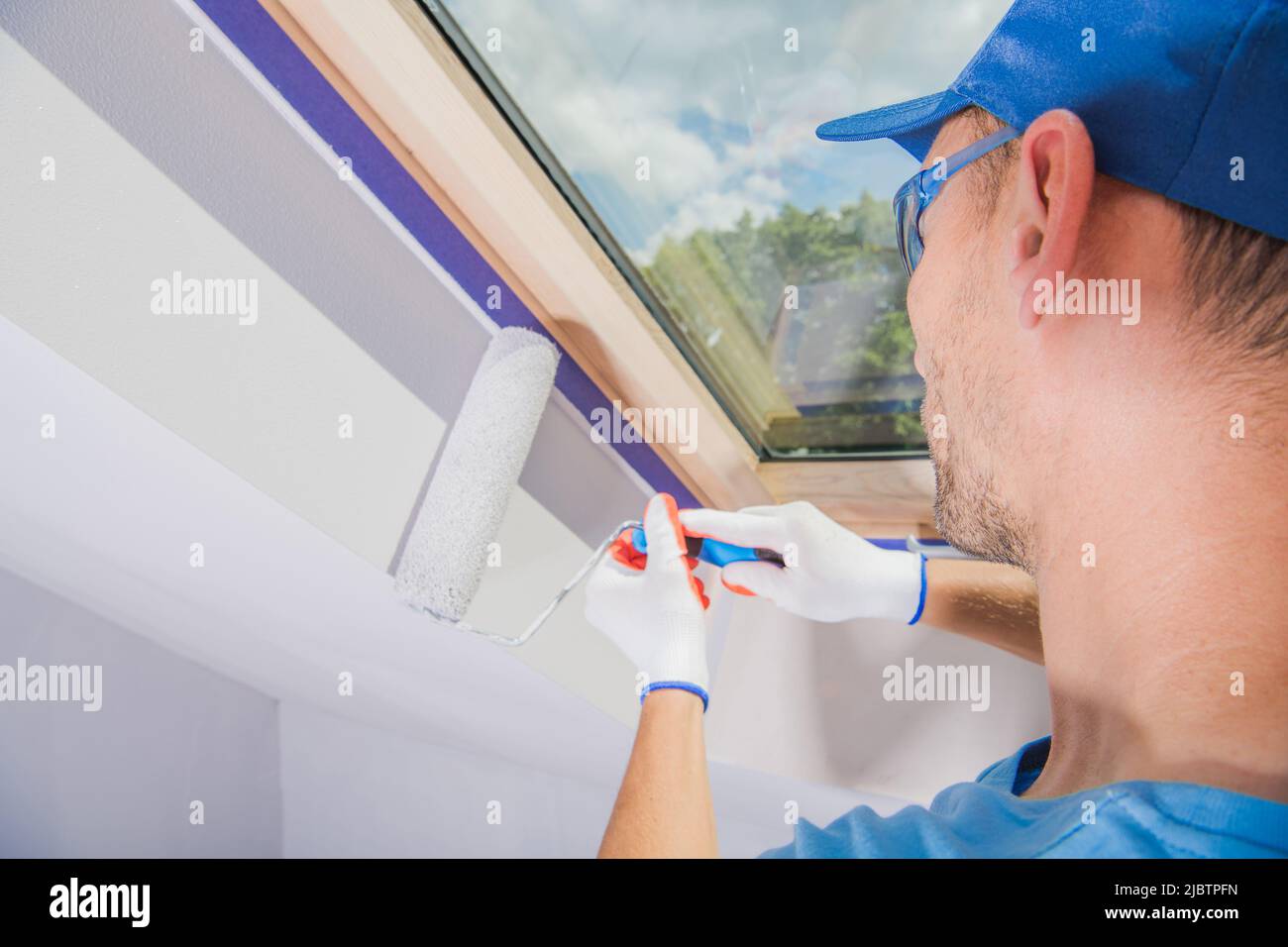Caucasian Construction Worker Pittura con cura le pareti con lo Skylight Window White dopo averlo fissato con nastro di pittura. Foto Stock