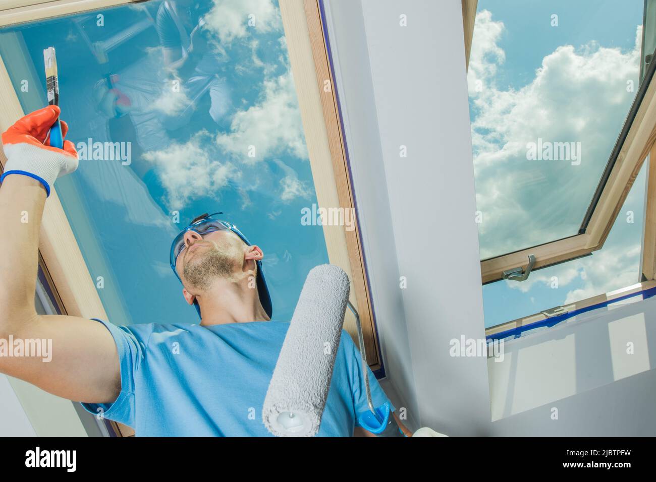 L'applicazione della vernice bianca con un piccolo pennello mentre tiene un rullo della vernice nell'altra mano. Il cielo blu brillante è visibile attraverso gli Skylights. Acceso Foto Stock