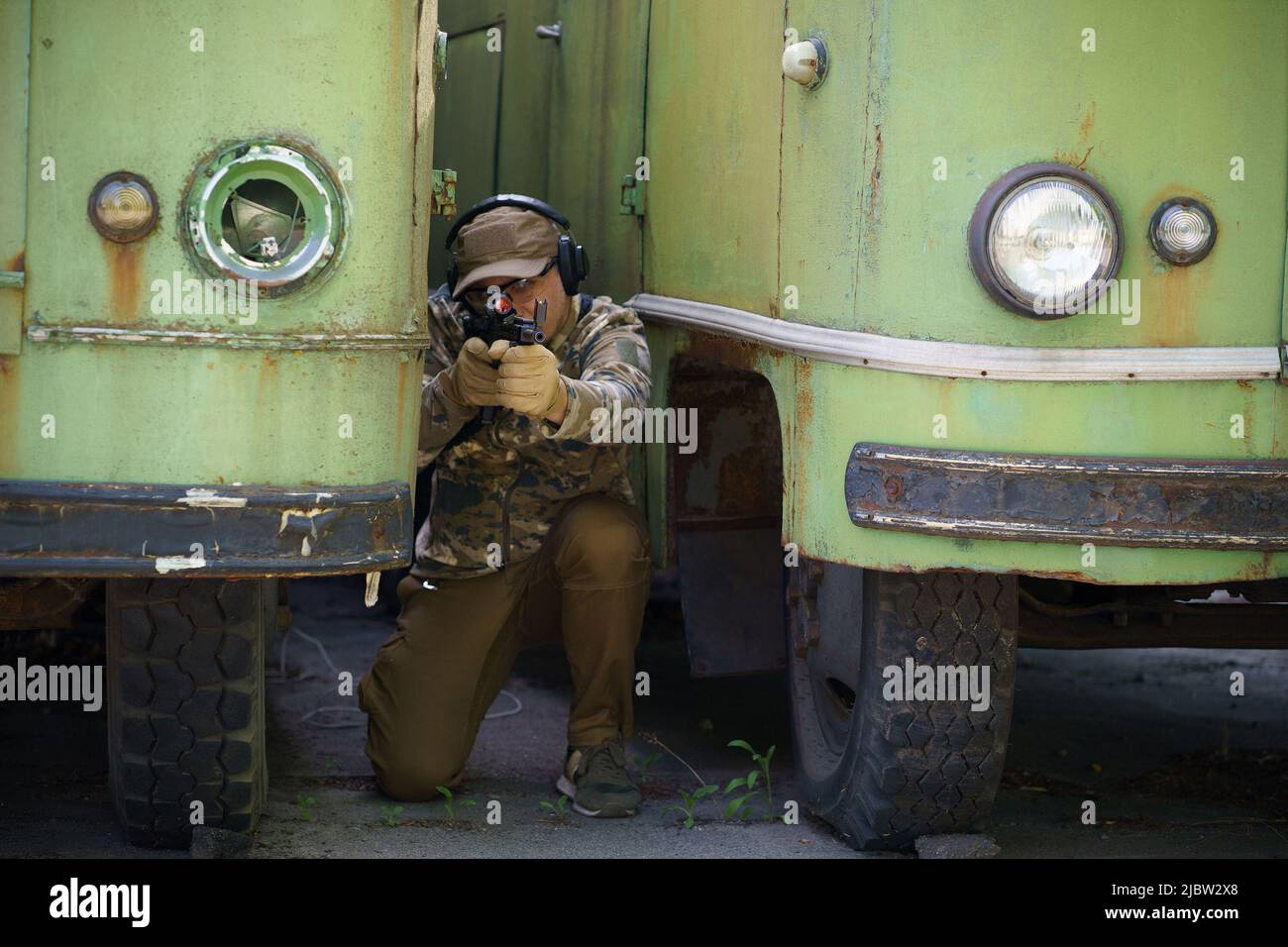 L'uomo militare si nasconde in posizione tra vecchi autobus arrugginiti con l'arma nelle mani. Sparatutto con pistola pronto a sparare. Ranger durante l'operazione militare. Combattimenti soldato di addestramento di forze speciali. Foto Stock