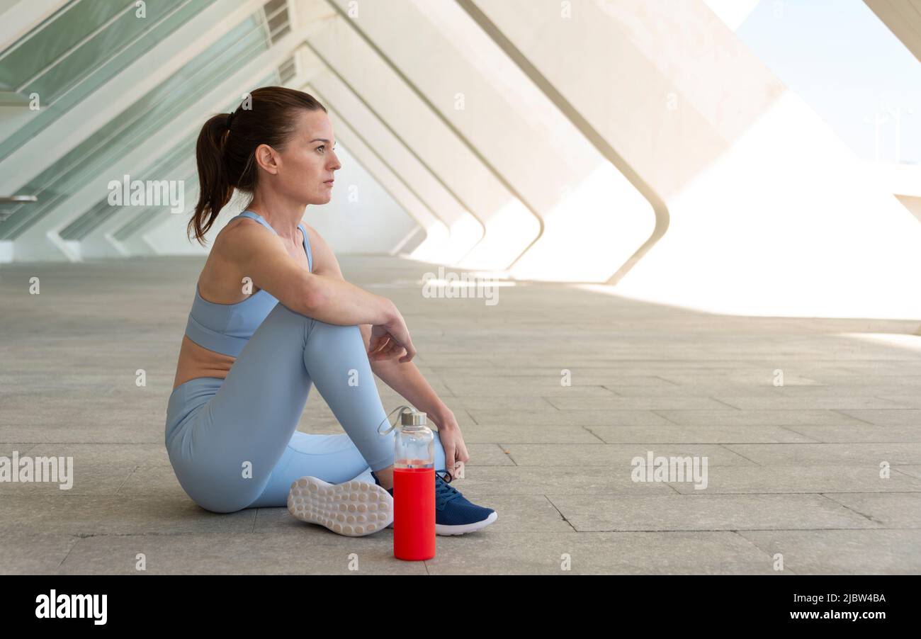 Donna sportiva e in forma seduta rilassante con una bottiglia d'acqua dopo l'esercizio, sfondo urbano. Foto Stock
