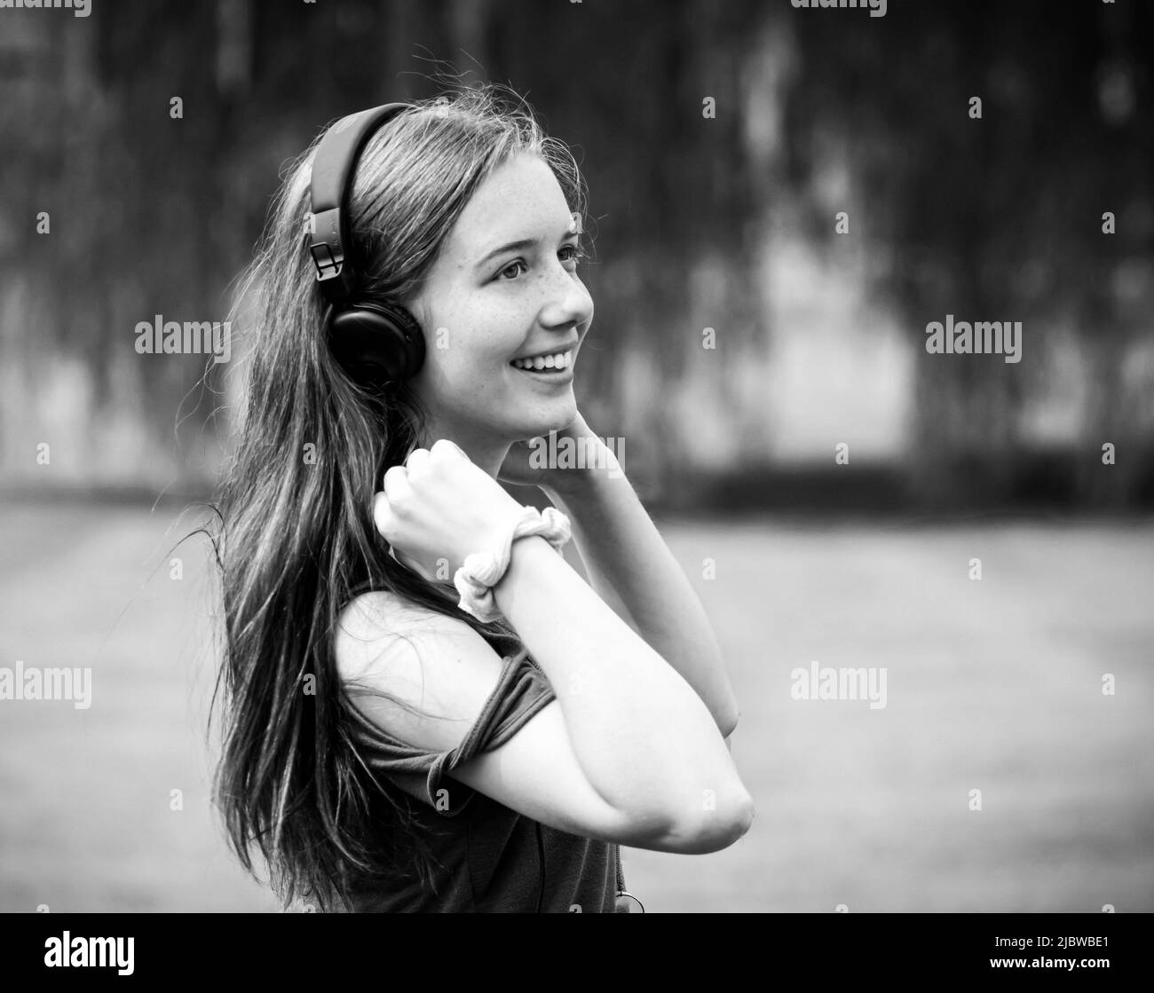 Giovane ragazza dai capelli lunghi con auricolari che sorridono su un prato d'erba con un albero di salice sullo sfondo durante l'estate in bianco e nero Foto Stock