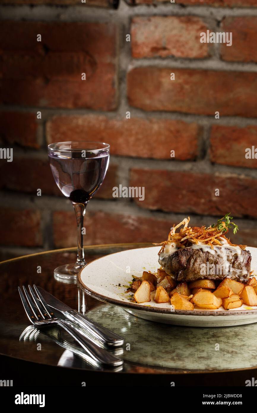 Bistecca alla griglia con patate dorate al forno servita con cipolla croccante sulla parte superiore e deliziosa bevanda su sfondo di mattoni. Ricetta deliziosa. Filetto di maiale Coo Foto Stock