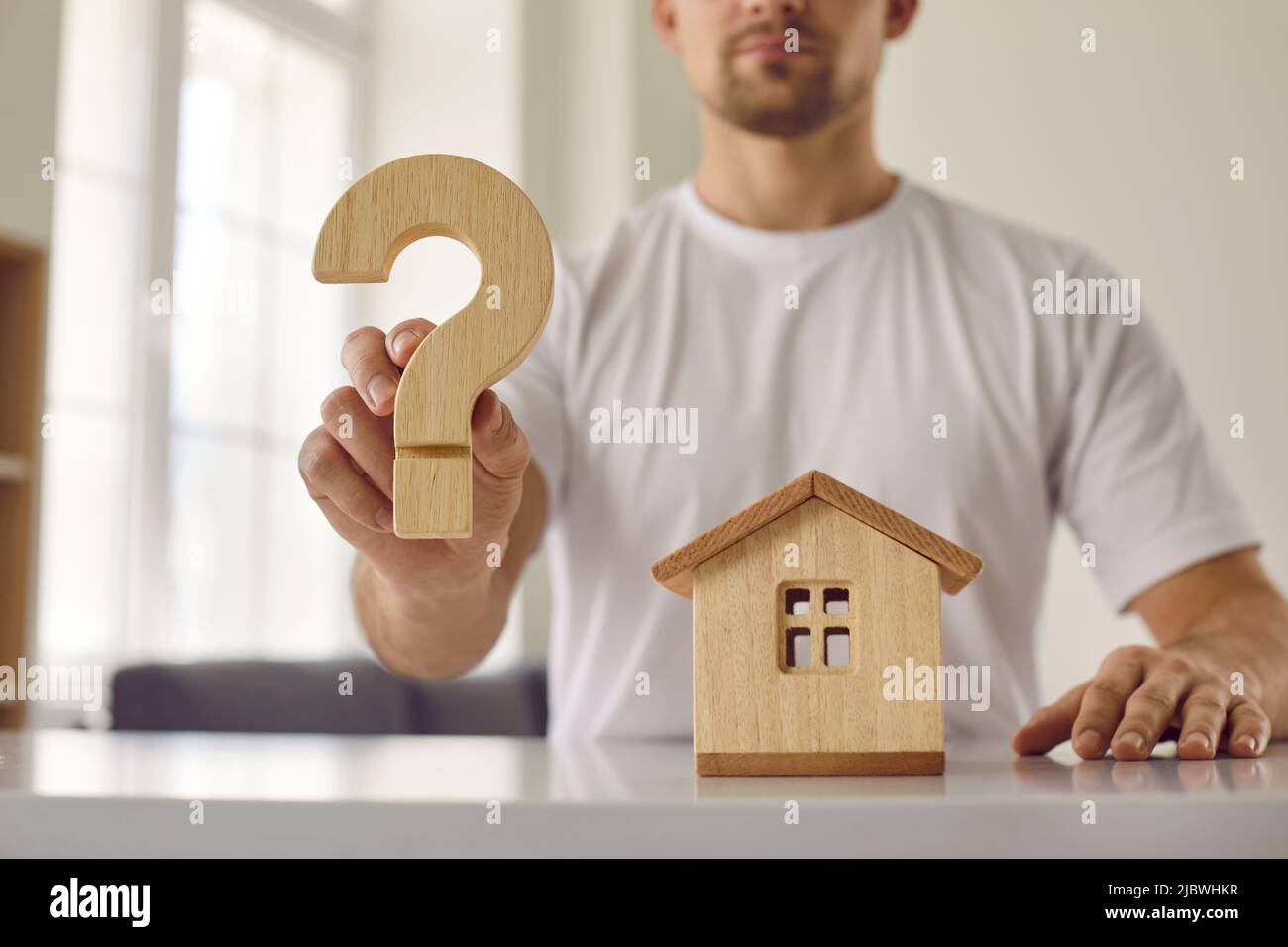 Confusione giovane uomo che è esitante circa l'acquisto della sua casa mostra legno punto di domanda. Foto Stock