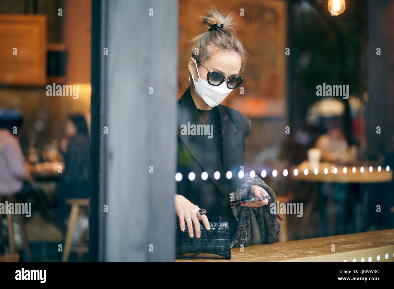 donna asiatica che indossa maschera in piedi accanto alla finestra in caffetteria guardando il telefono cellulare Foto Stock