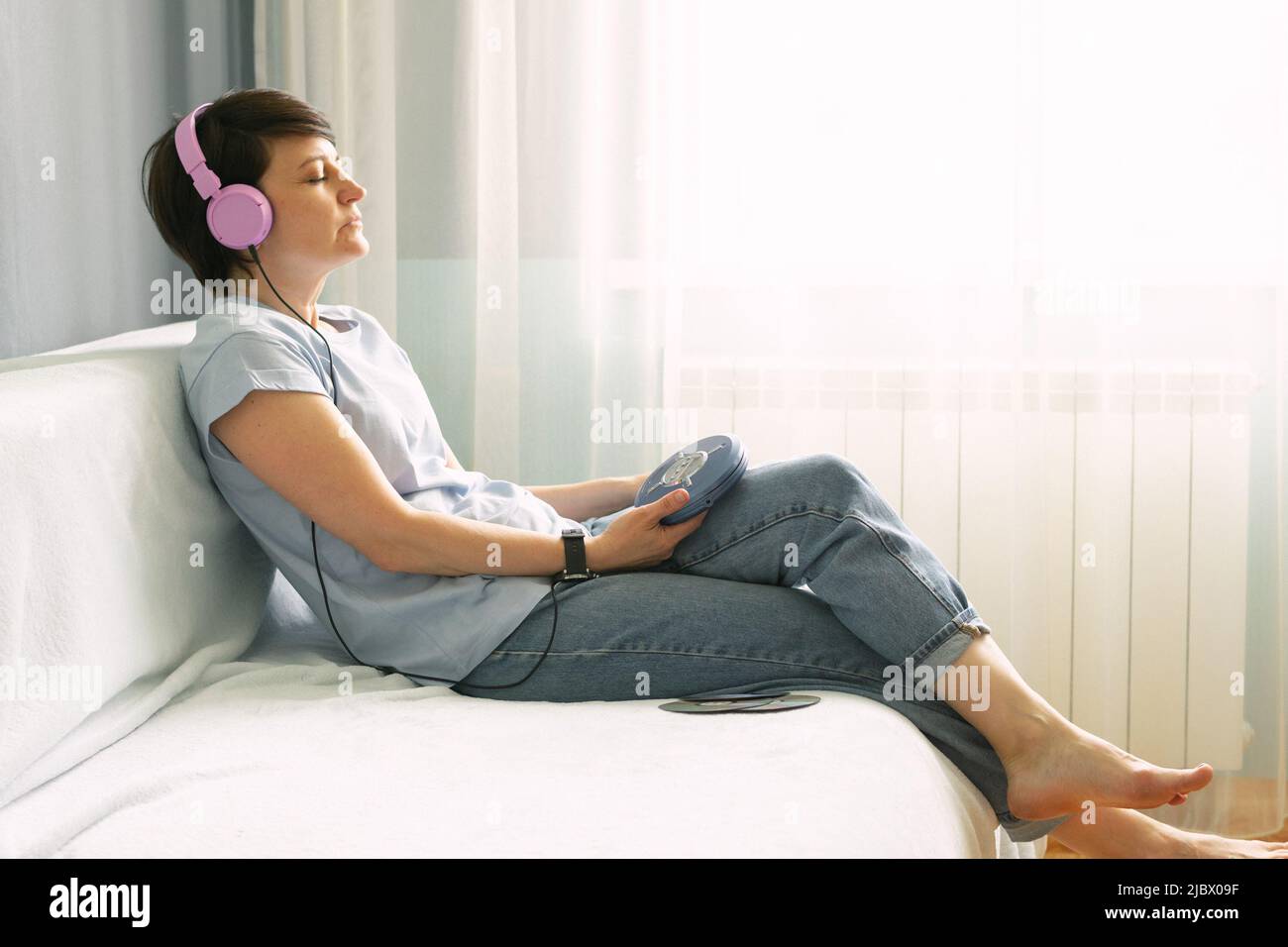 Una donna adulta in cuffie rosa si siede sul divano a casa e ascolta la musica sui lettori CD Foto Stock