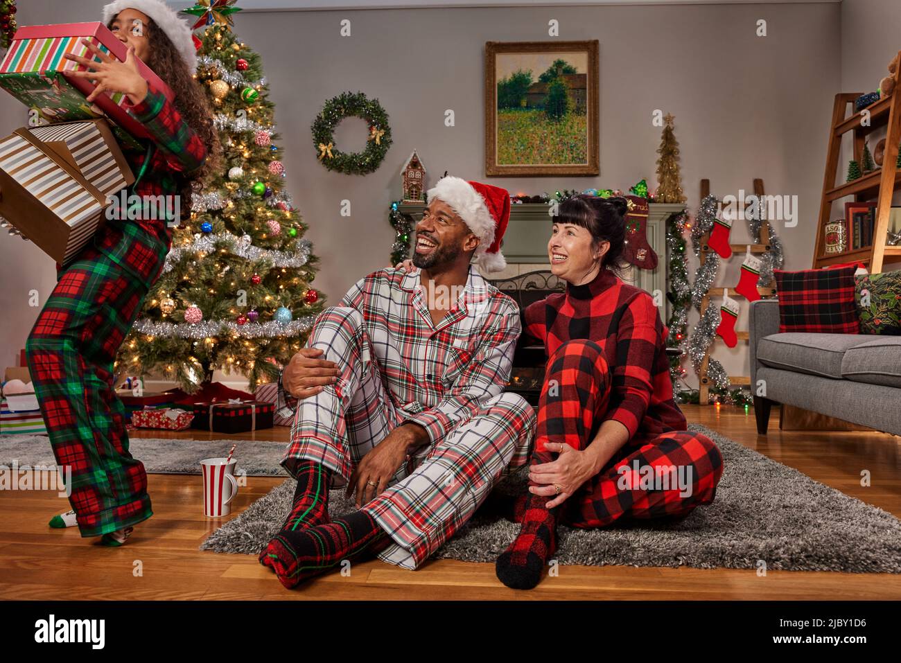 Vista frontale di una coppia mista gara con la loro giovane figlia a Natale, con cappelli Babbo Natale seduti insieme sul pavimento, regali di apertura. Foto Stock