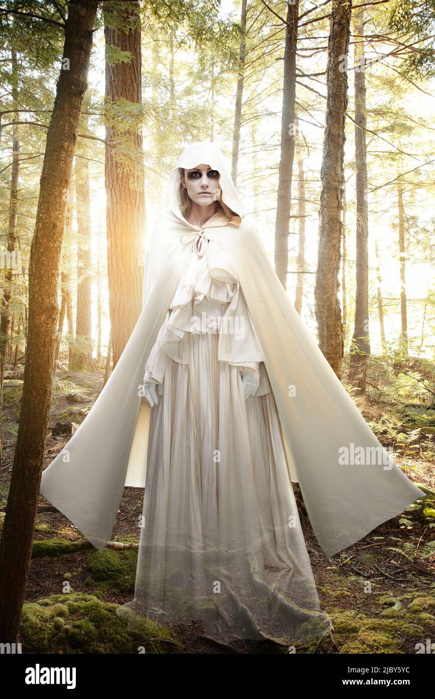 Fantasma femminile con cerchi neri intorno ai suoi occhi, vestito tutto in bianco in piedi in una foresta all'alba Foto Stock