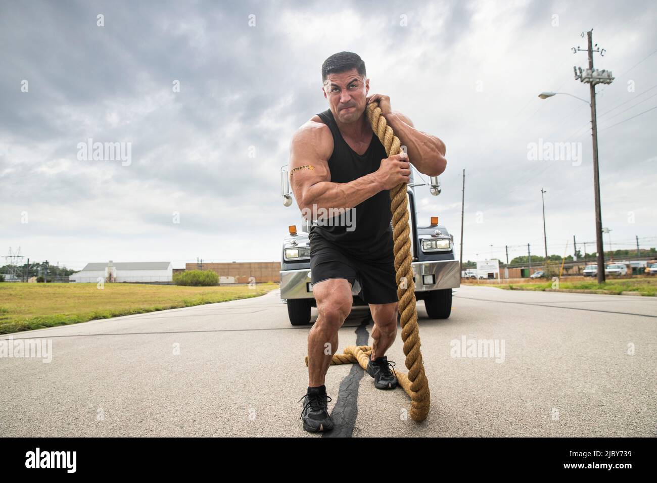 Uomo ispanico muscoloso che indossa la canotta con un tatuaggio "forte" sulla sua bicicletta, traendo il semi-camion con una grande fune di traino Foto Stock