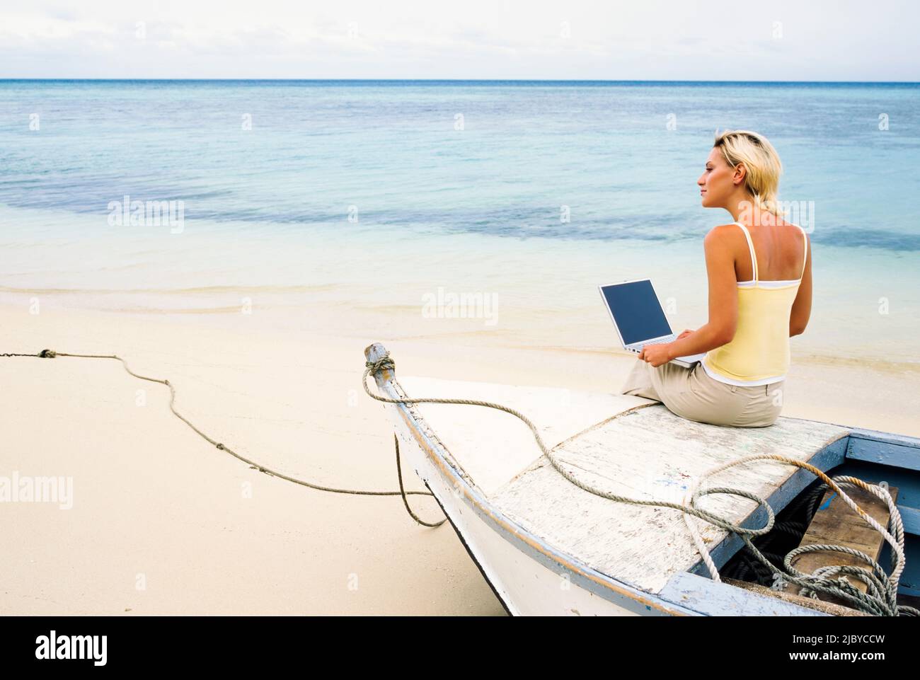 Giovane donna seduta sull'arco di una piccola barca con il suo computer portatile su una spiaggia tropicale Foto Stock