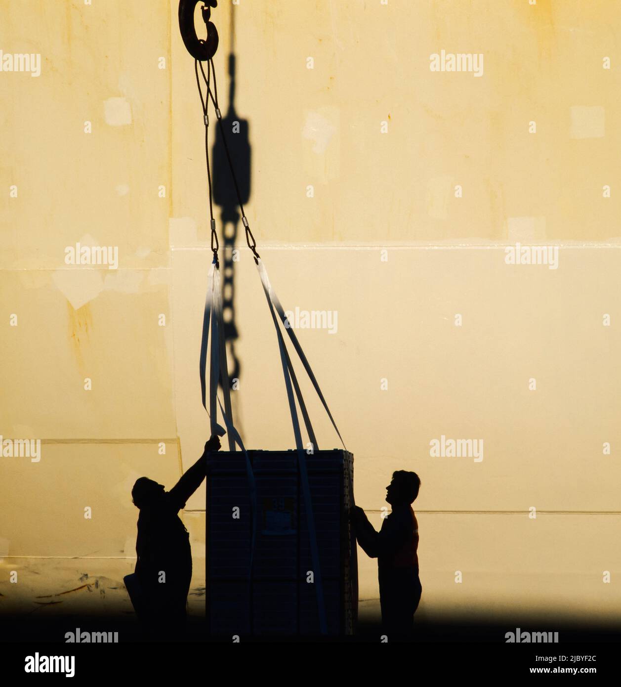 Silhouette di due uomini che guidano il carico fissato al grande gancio di fronte allo scafo delle navi Foto Stock