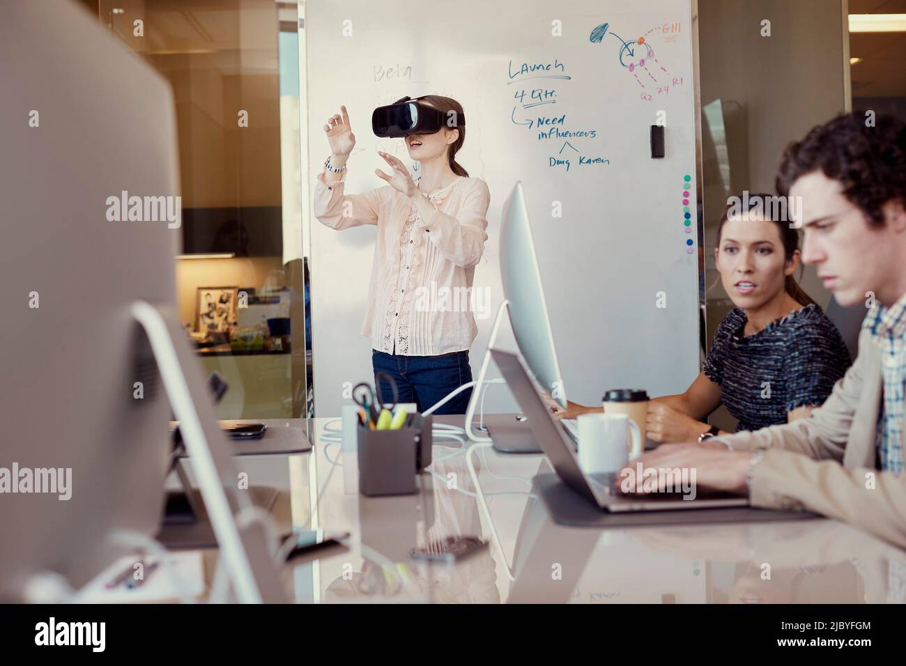 -Lavoratori che lavorano al computer in ufficio mentre la donna utilizza Virtual Reality Headset in piedi di fronte alla lavagna di cancellazione a secco con diagramma di flusso in background Foto Stock