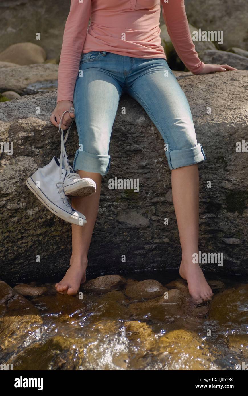 Giovane donna che tiene scarpe seduti su roccia e piedi per pagaiare nel fiume Foto Stock