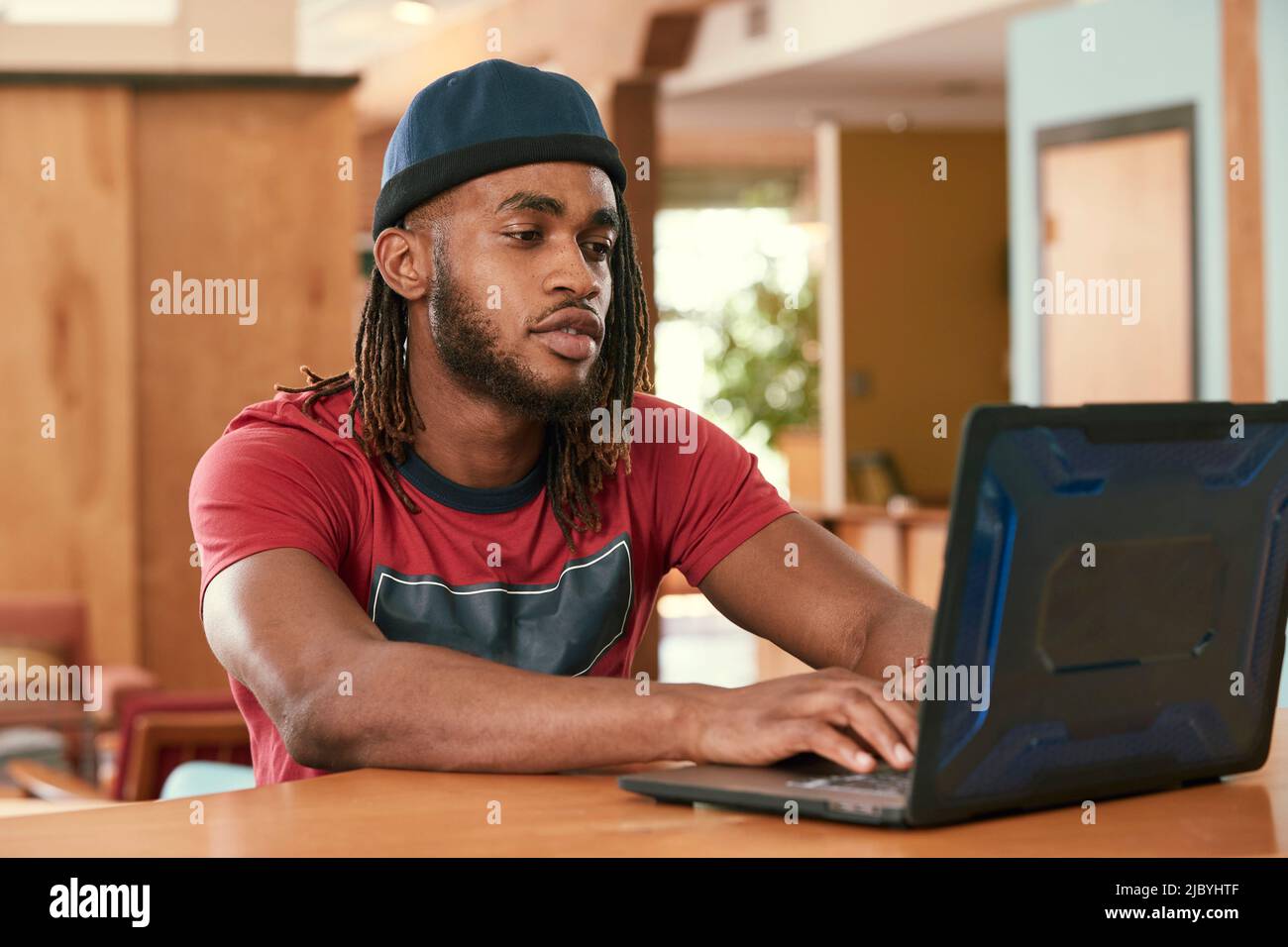 Ritratto di giovane uomo etnico con maglietta rossa e cappello a maglia , seduto al bar in cucina del centro loft con computer portatile Foto Stock