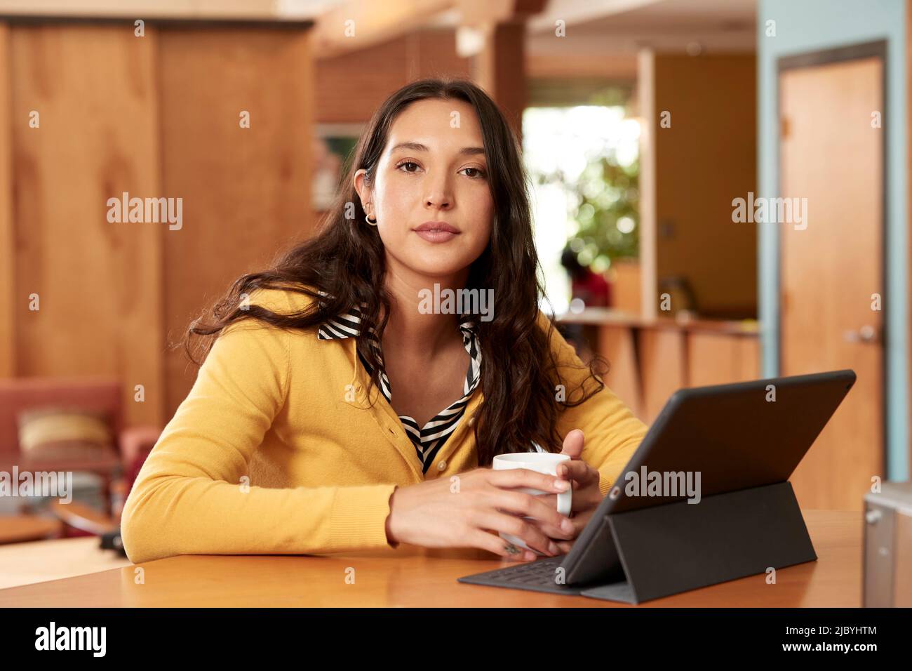 Di giovane etnia donna che indossa maglione giallo con blusa a righe bianche e nere, seduto al bar in cucina del centro loft con iPad e tazza di caffè Foto Stock