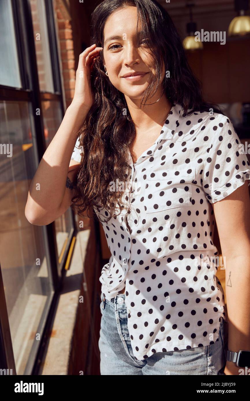 ritratto di giovane etnia donna indossando polka puntino blusa e tagliare jean shorts in piedi da banca di finestre in centro loft Foto Stock