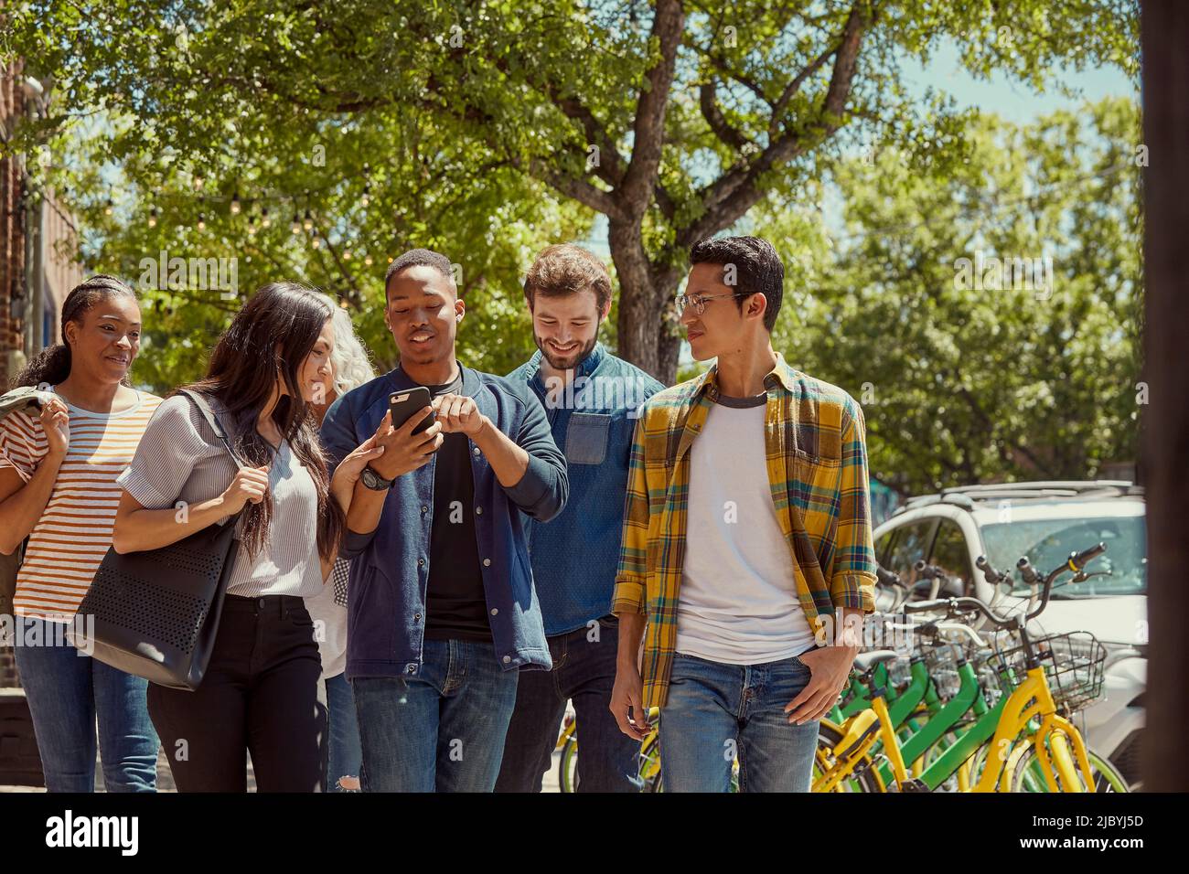 Gruppo di giovani collaboratori che camminano lungo il marciapiede insieme, giovane uomo che mostra agli amici qualcosa sul cellulare Foto Stock