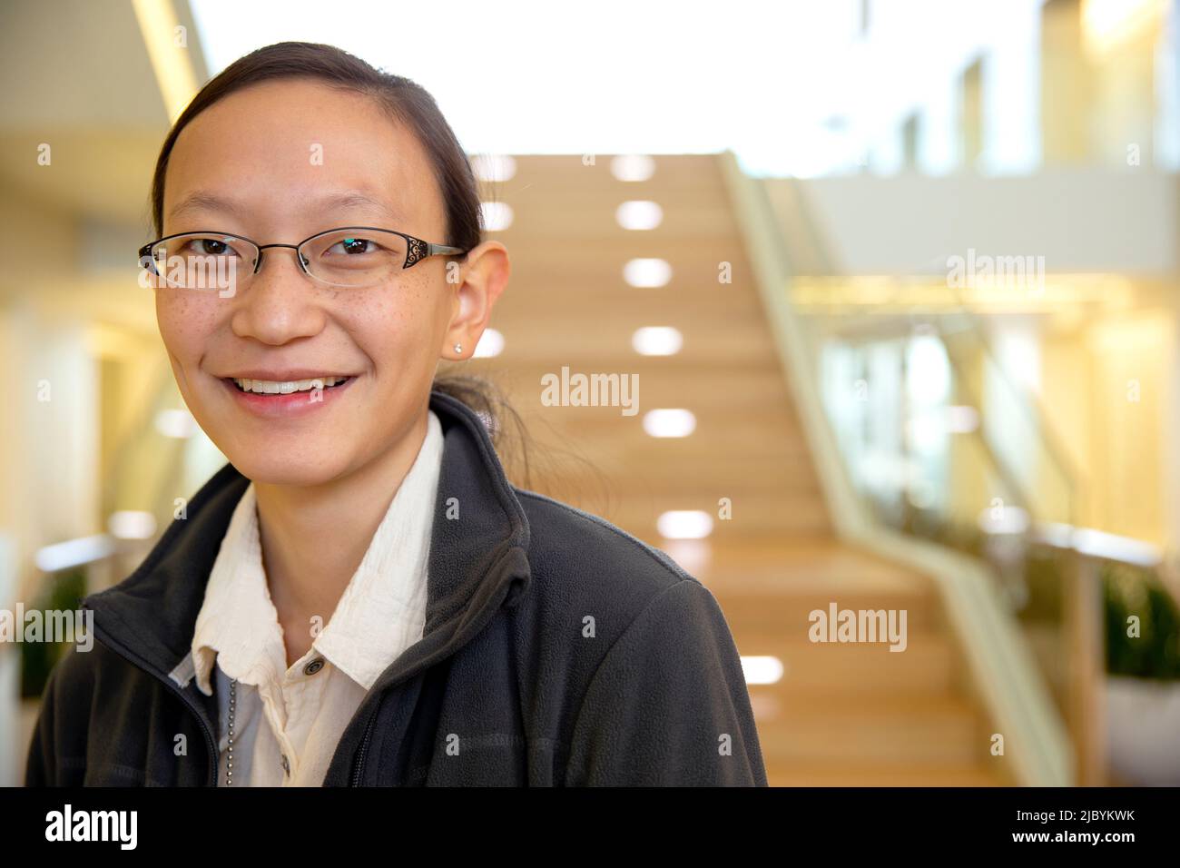 Ritratto di giovane etnia donna che indossa occhiali in piedi dalla scala nella lobby dell'edificio, sorridente guardando verso la macchina fotografica Foto Stock
