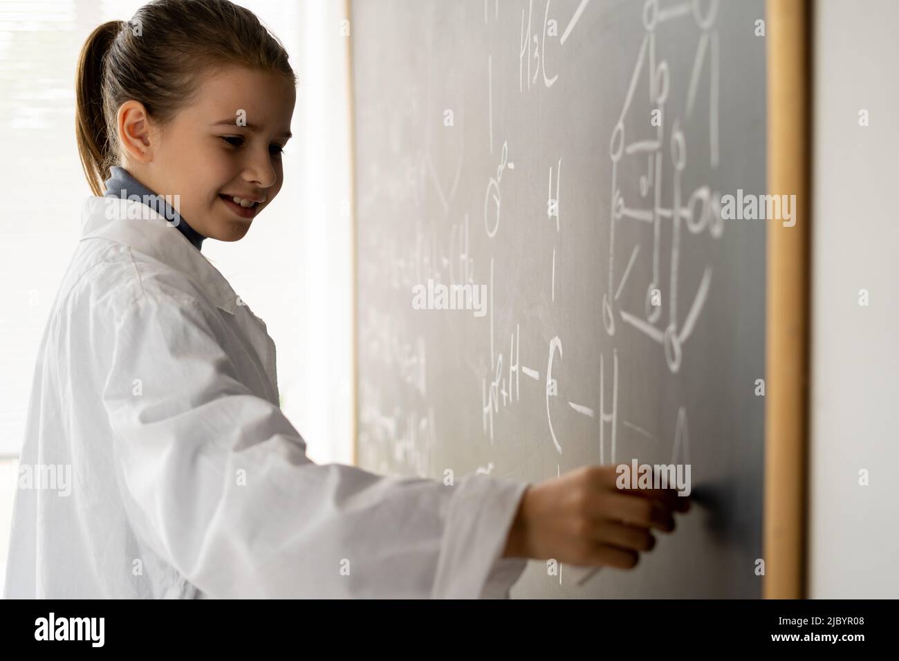 sorridendo femmina studente college scrittura su lavagna durante la lezione di chimica, scolgirl in bianco cappotto scrittura formule con gesso per esame di chimica Foto Stock