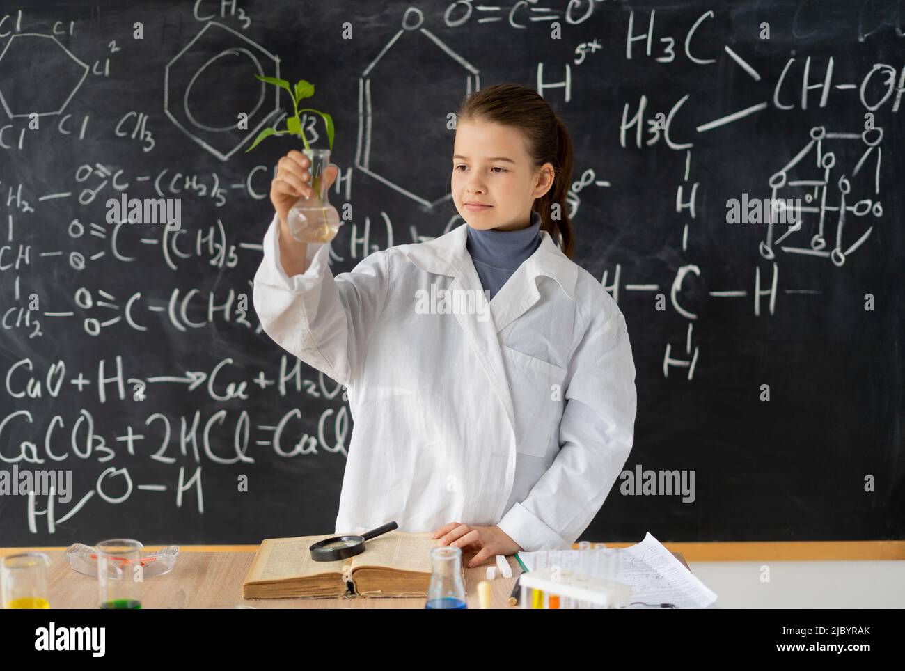 Piccolo biochimico toccando provetta con pianta piccola, ragazza scienziata presentare matraccio con pianta all'interno, sorriso e guardare a macchina fotografica con altri strumenti in Foto Stock