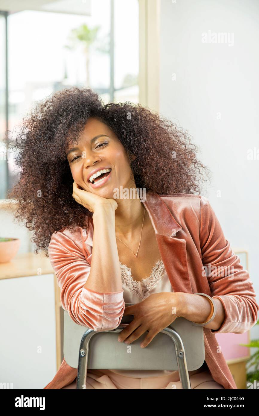 Colpo medio di una corsa mista, donna di mezza età che si appoggia sulla schiena di una sedia ridendo. Foto Stock